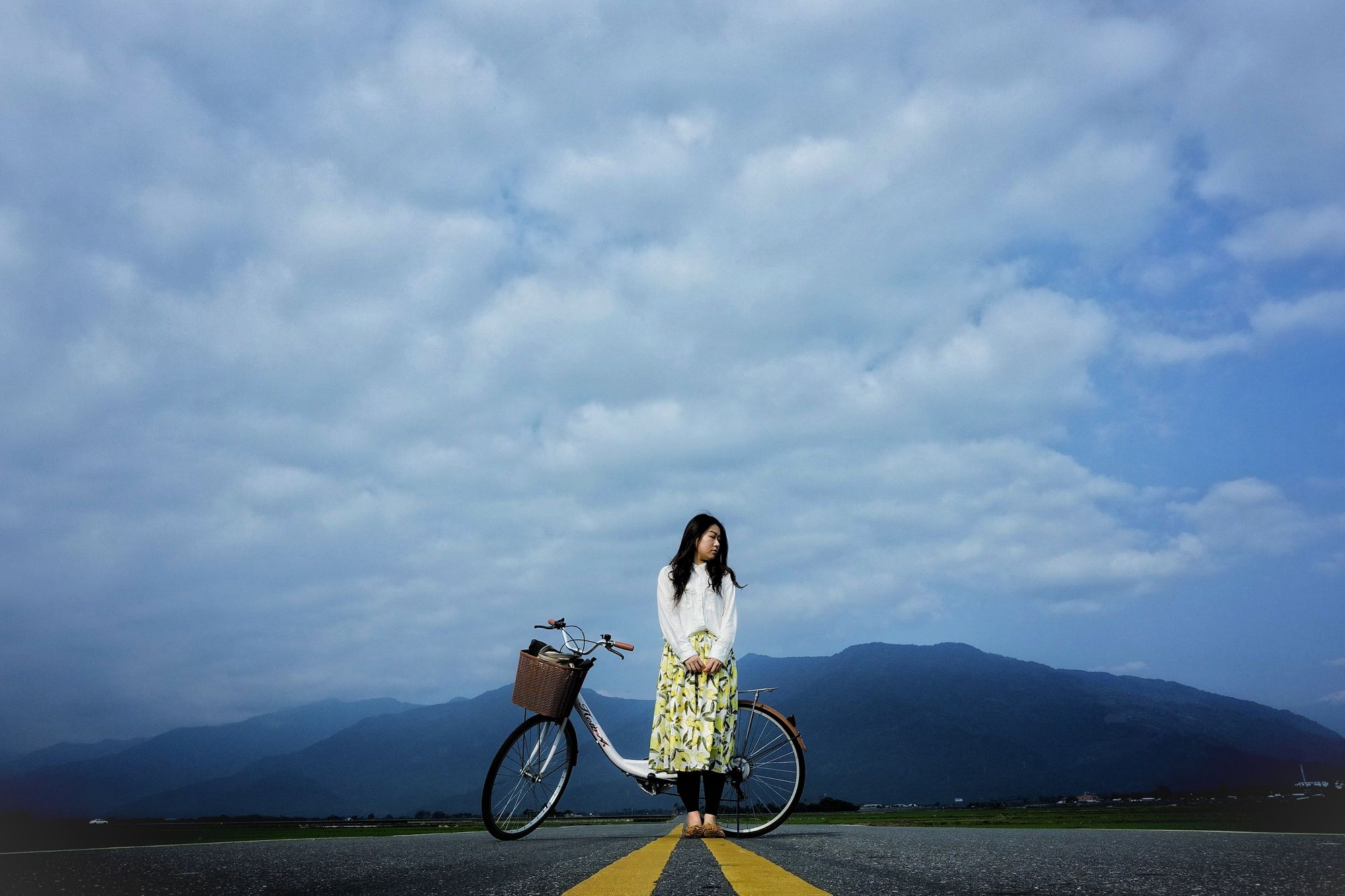 Woman waiting on an empty street with her bicycle, low hills in the background.